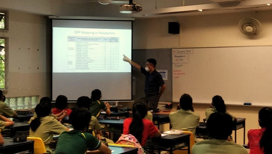 A masked teacher points at a projected slide titled "DPP Mapping to Polytechnic" while students sit at desks.